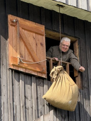Gast sieht durch ein Luuk aus der Mühle Ein älterer Mann lehnt aus einem Fenster und zieht einen großen Sack nach oben.