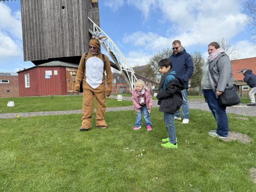 Kinder beim Eiertrullern Person im Tierkostüm mit Kindern und Erwachsenen in einer Wiese vor einer Mühle.