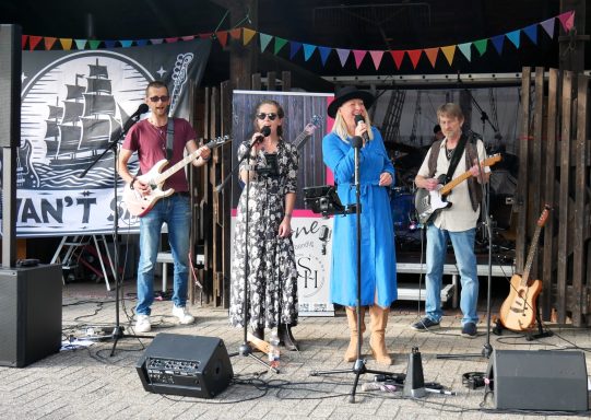 Band spielt live auf einer Bühne, mit Sängerin und Bassist in buntem Ambiente.