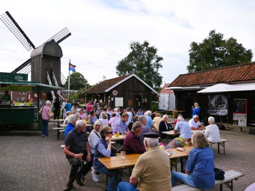 Menschen sitzen an Tischen im Freien, umgeben von Gebäuden und einer Windmühle.