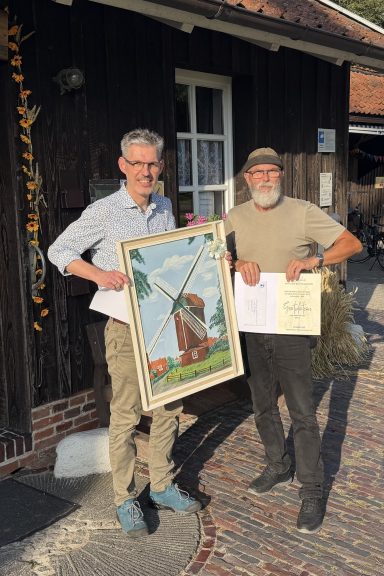 Zwei Männer stehen vor einem Gebäude und halten ein Gemälde sowie Zertifikate.