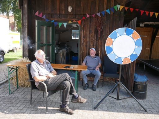 Zwei Männer sitzen entspannt vor einem Holzschuppen, ein Glücksrad steht daneben.