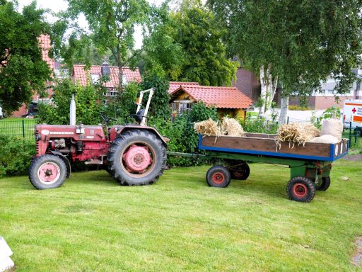 Roter Traktor neben einem Wagen mit Heuballen, umgeben von Bäumen und Grünflächen.