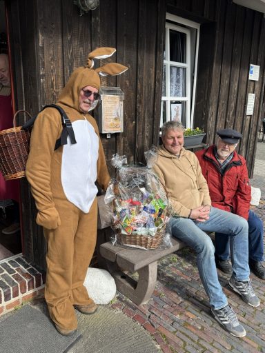 Osterhase Wilhelm mit dem erknobelten Präsentkorb Mann im Hasenkostüm neben zwei Personen vor einem Holzhaus mit einem Korb voller Süßigkeiten.