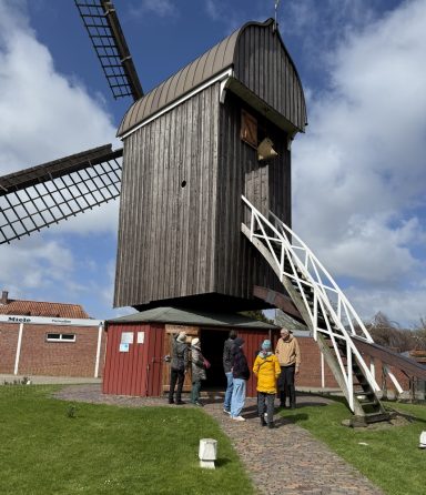 Roland bei einer Führung Eine Holzwindmühle mit Besuchern auf dem Weg zum Eingang und blauem Himmel.